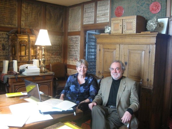 a woman and a man sit together indoors in front of a computer and a pile of papers